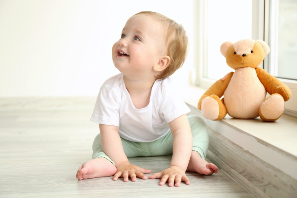 Ein kleiner, blonder Junge sitzt neben einem Teddybären und lächelt.