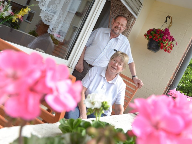 Ein Rentner-Ehepaar posiert hinter pinken Geranien auf dem Balkon für ein Foto.