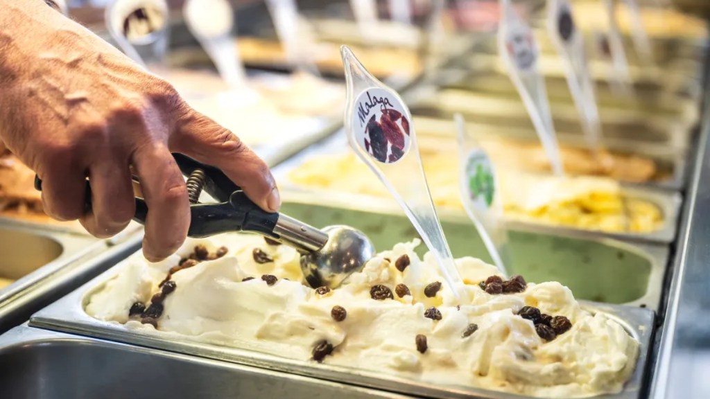 Eisverkäufer entnimmt eine Kugel Eis der Sorte „Malaga“.