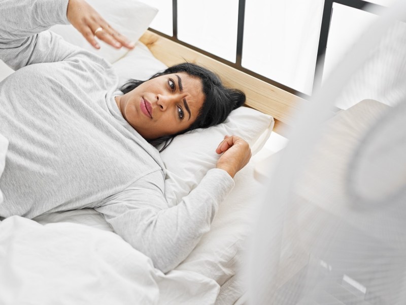 Eine Frau liegt in einem Bett mit Sommer-Bettwäsche und fächert sich kühle Luft zu.