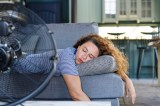 Ein Foto zeigt eine junge Frau beim Schlafen bei Hitze auf einer Couch, die sich mit einem Ventilator abkühlt.