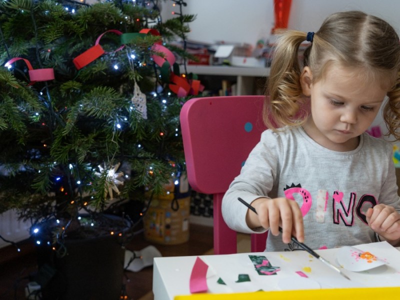 nützliche Geschenke Kinder: ein kleines Mädchen sitzt neben einem Weihnachtsbaum am Tisch und bastelt