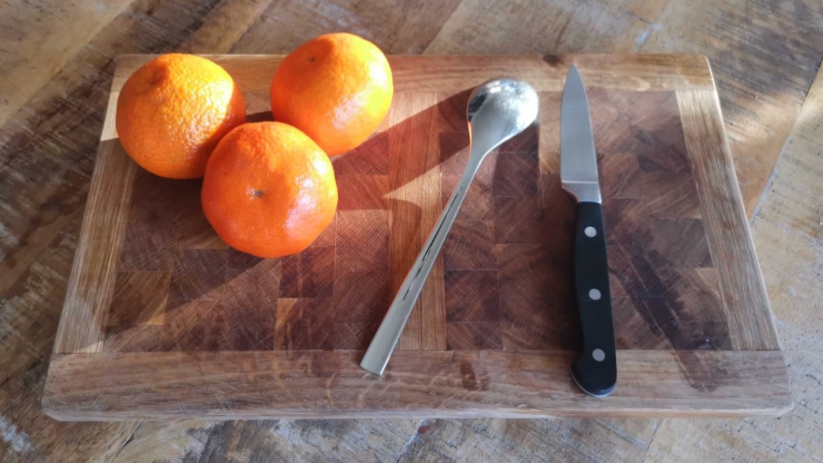 Mandarinen schälen: drei Mandarinen, ein Esslöffel und ein Messer liegen auf einem Holzbrett