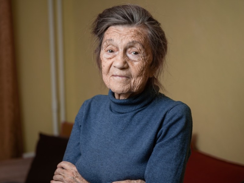 Eine alte Frau hat ihre Hände vor dem Bauch gekreuzt und schaut neutral in die Kamera.