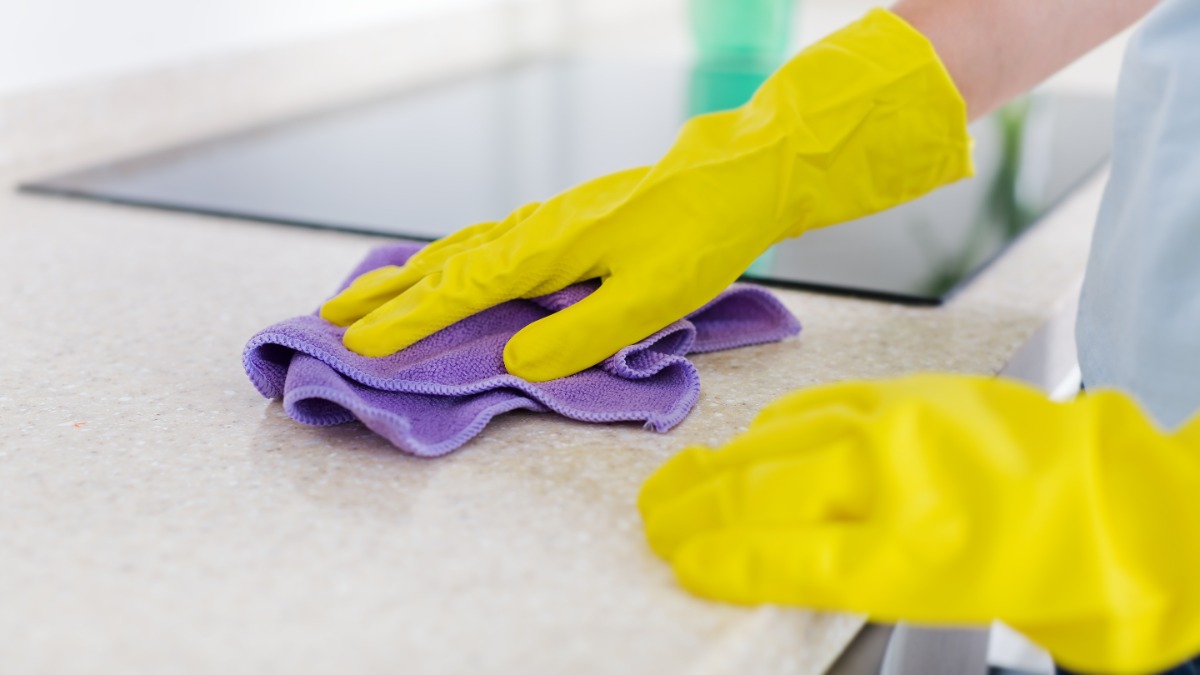eine Frau mit gelben Putzhandschuhen reinigt eine Küchenarbeitsplatte mit einem lila Schwammtuch