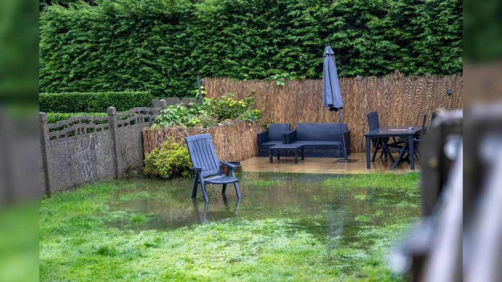 Eine große Pfütze auf einer Rasenfläche im Garten, in der Gartenmöbel stehen.
