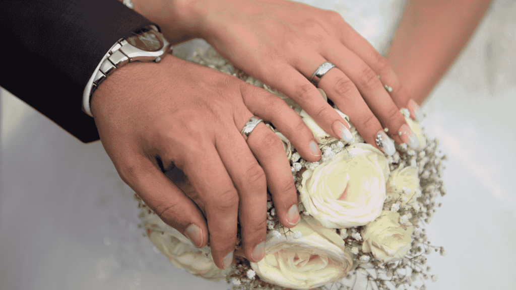 Hochzeitstage Namen, zwei Hände mit Eheringen liegen auf einem cremefarbenen Blumenstrauß
