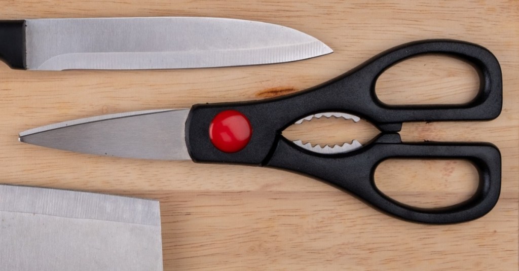 eine Schere liegt auf einem Holzbrett neben zwei Messern
