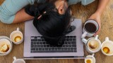 Eine Frau umgeben von leeren Tassen, die müde mit dem Kopf auf der Tastatur eines Laptops liegt.