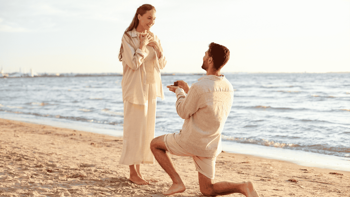heiratsantrag ideen, Mann kniet vor Frau im Sand und hält ihr eine Schmuckschachtel entgegen