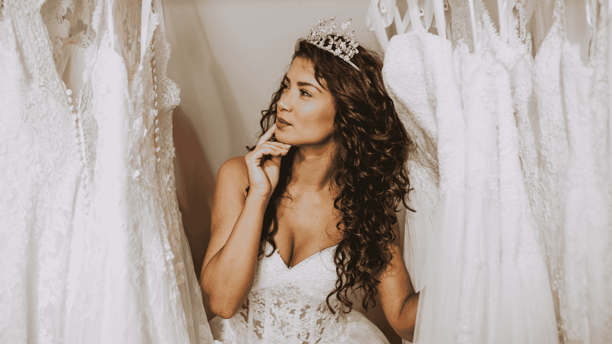 Hochzeitskleid kaufen, Braut steht zwischen Hochzeitskleidern und guckt fragend