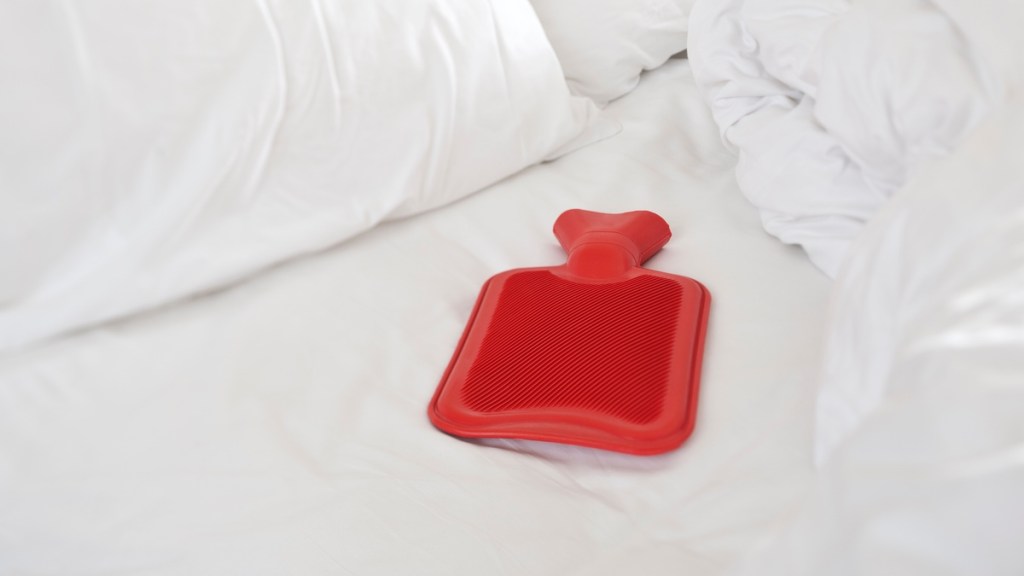 Rote Wärmflasche liegt in einem Bett.